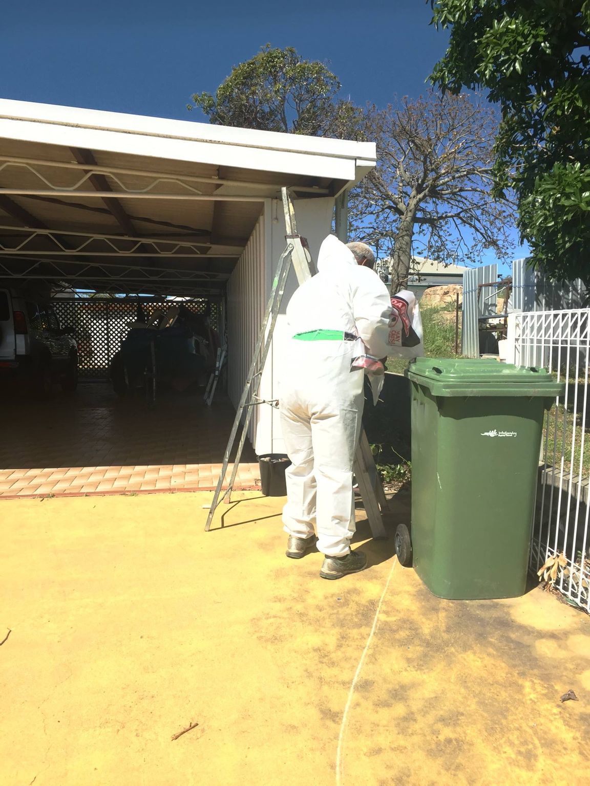 Person in white protective suit by green trash bin, near carport.
