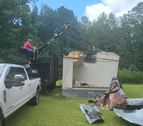 A man is sitting on top of a truck while a crane removes a shed.