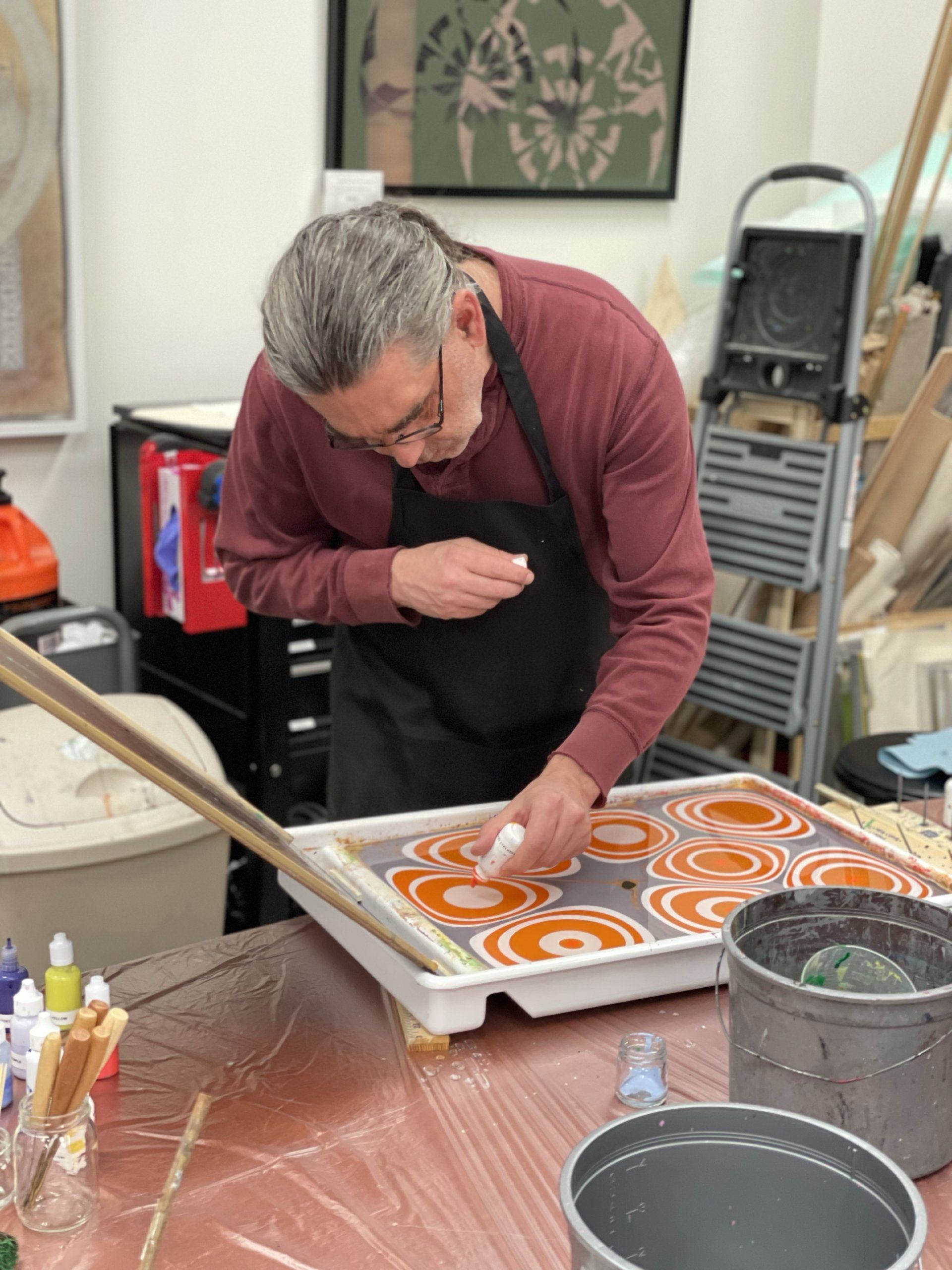 Man applies color to his marbling bath in orange and white on a gray field.