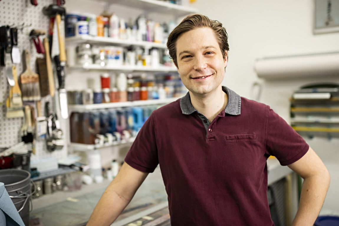 Artist Garubba stands in front of tool rack and paint shelves of studio.