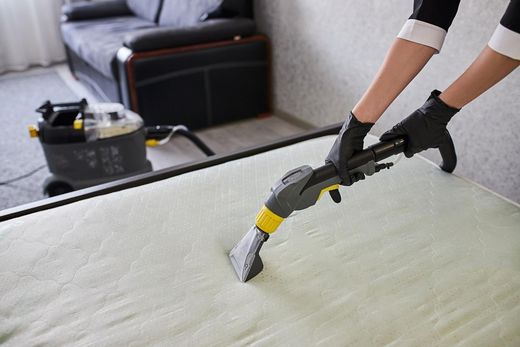 Person in black gloves vacuums a mattress with a cleaning machine in a room, sofa in background.