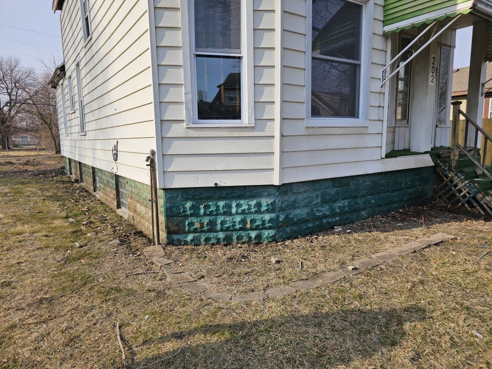 A white house with green trim and a brick foundation, in a yard with sparse grass.