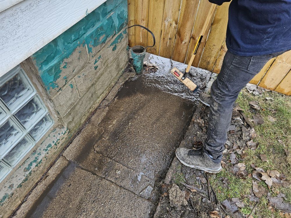 Person cleaning concrete near a building, wearing dark jeans and gray shoes. Beside a wooden fence.