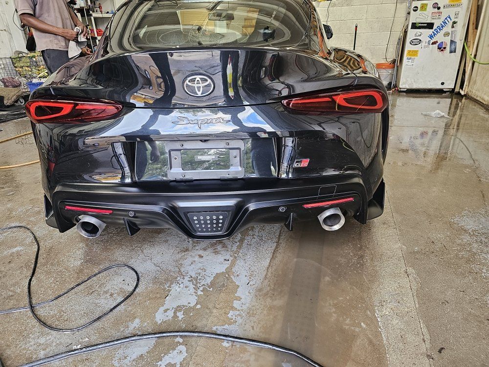 Black Toyota Supra sports car, rear view, in a garage with water on the floor, person standing nearby.