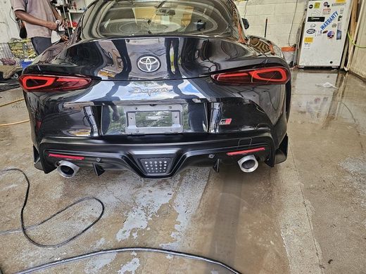 Black Toyota Supra sports car, rear view, in a garage with water on the floor, person standing nearby.