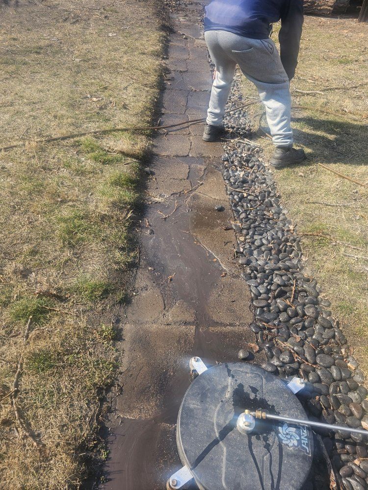 Person power washing a paved pathway next to a rock border, on a grassy lawn.