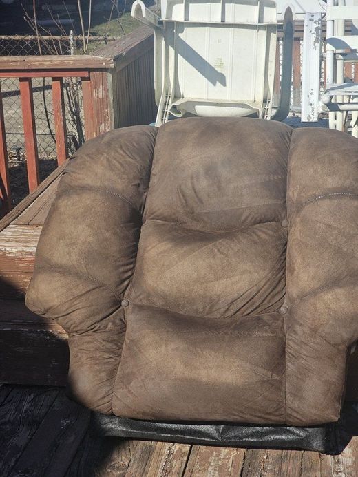 Brown, worn recliner on an outdoor wooden deck, with a white chair in the background.