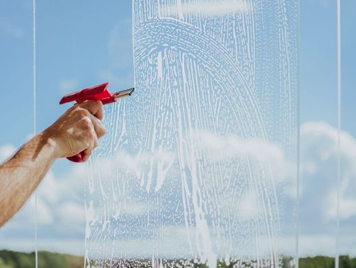 Hand spraying soapy solution onto a window against a blue sky with clouds.