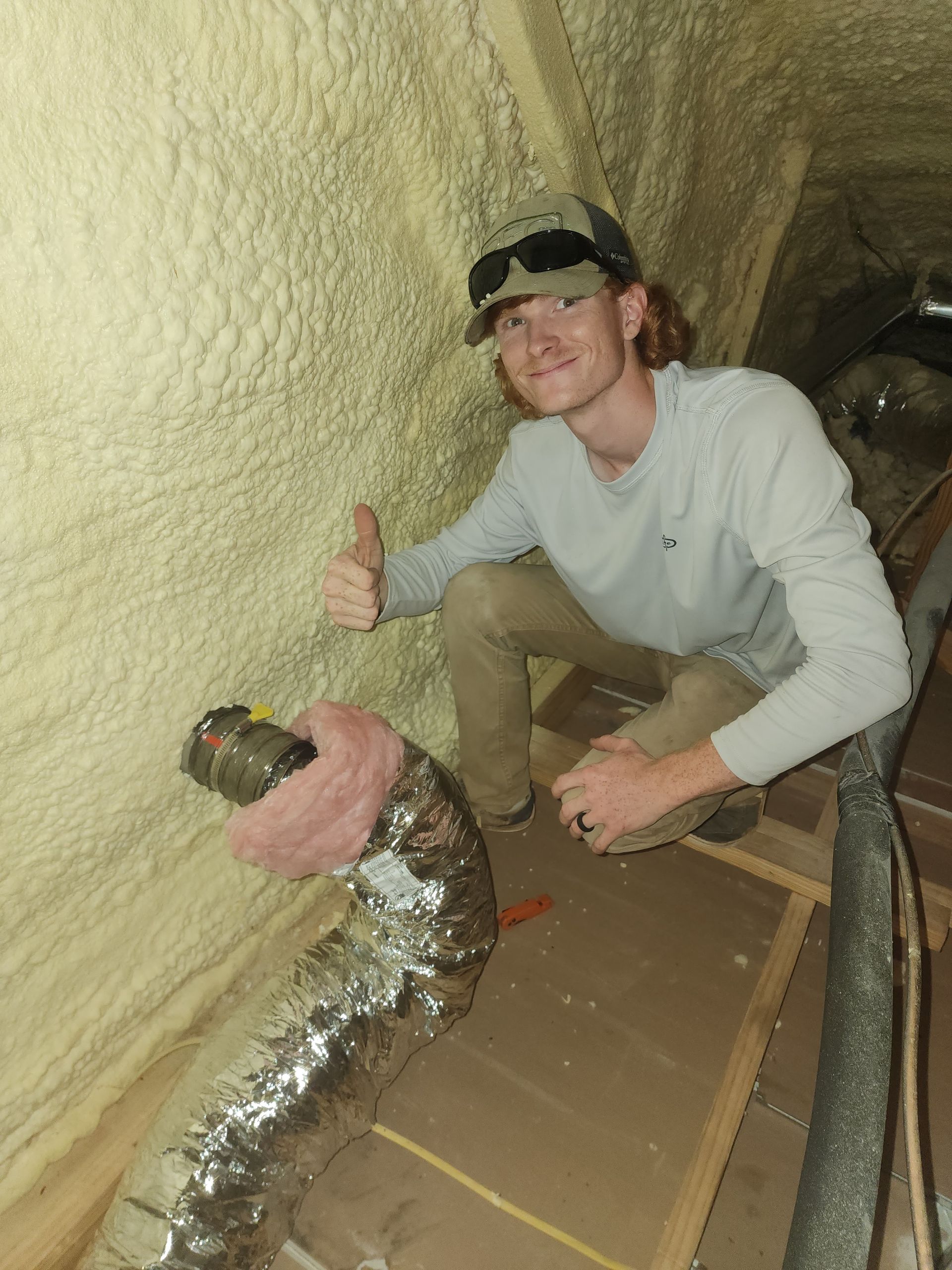 Man in a hat giving a thumbs-up inside a crawl space insulated with spray foam, near ductwork.
