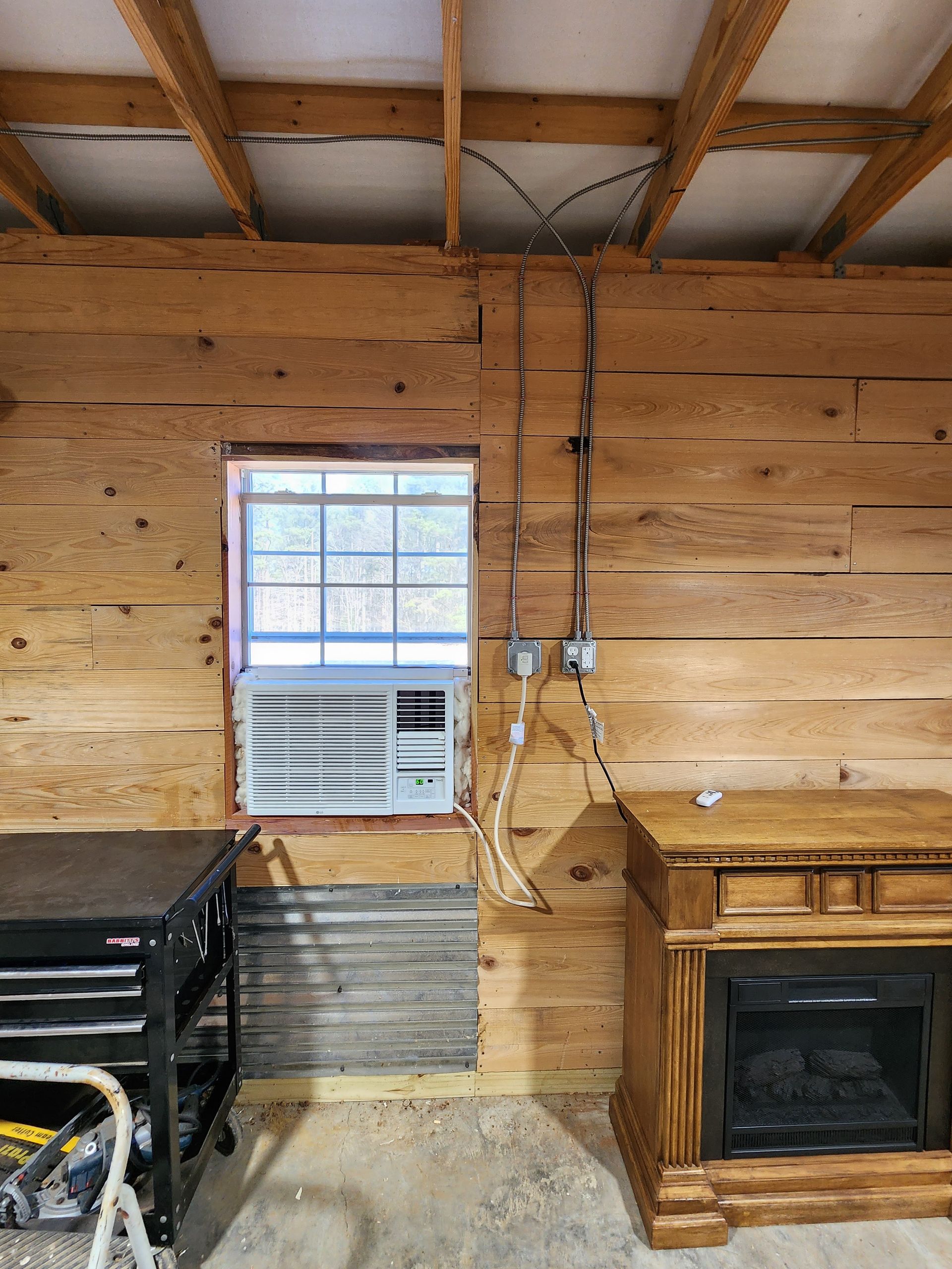Wood-paneled room with a window, air conditioner, fireplace, and tool chest. Electrical wiring visible on the wall.