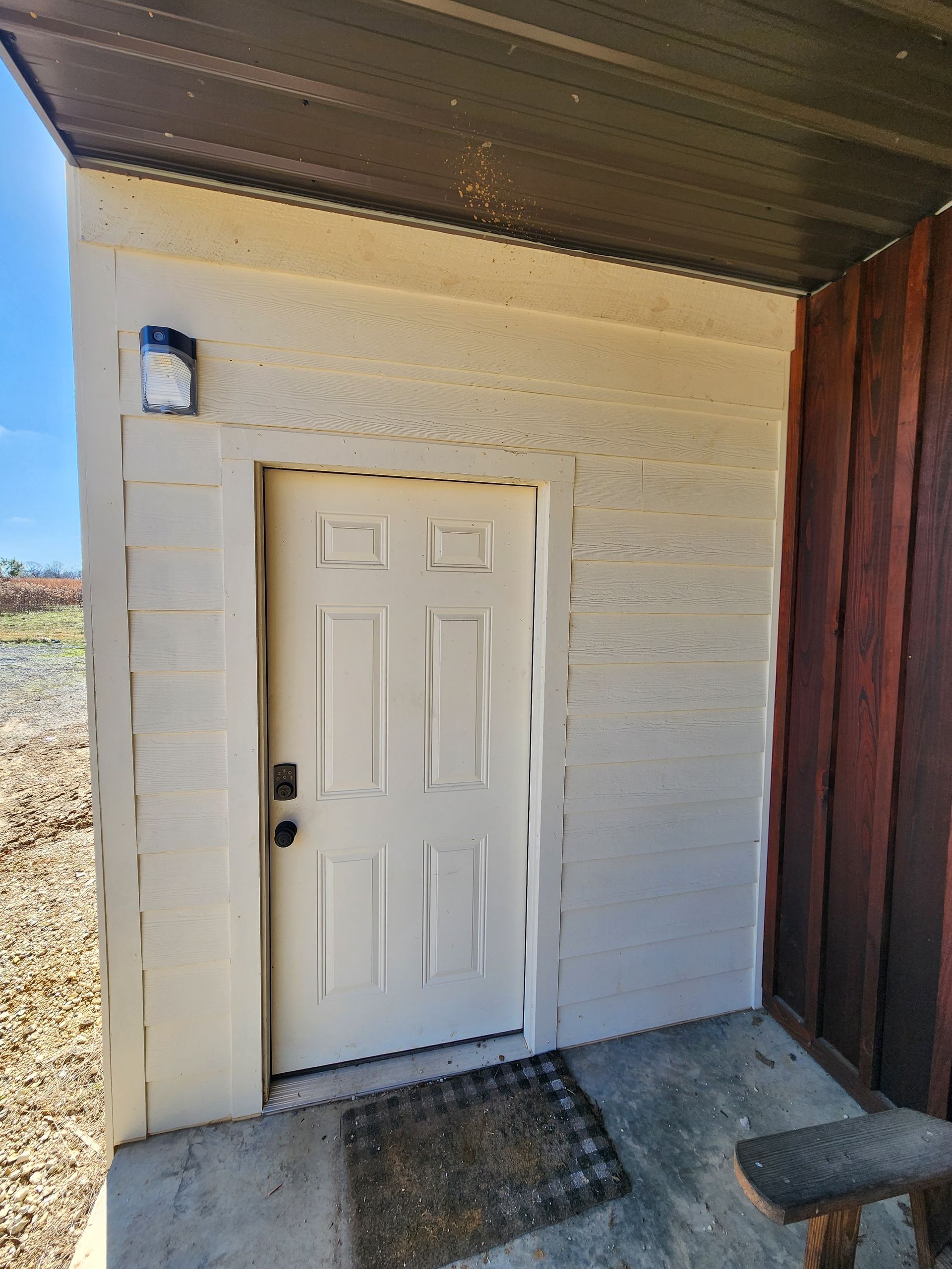 Beige door with black handle and small welcome mat on a concrete porch under a dark roof.
