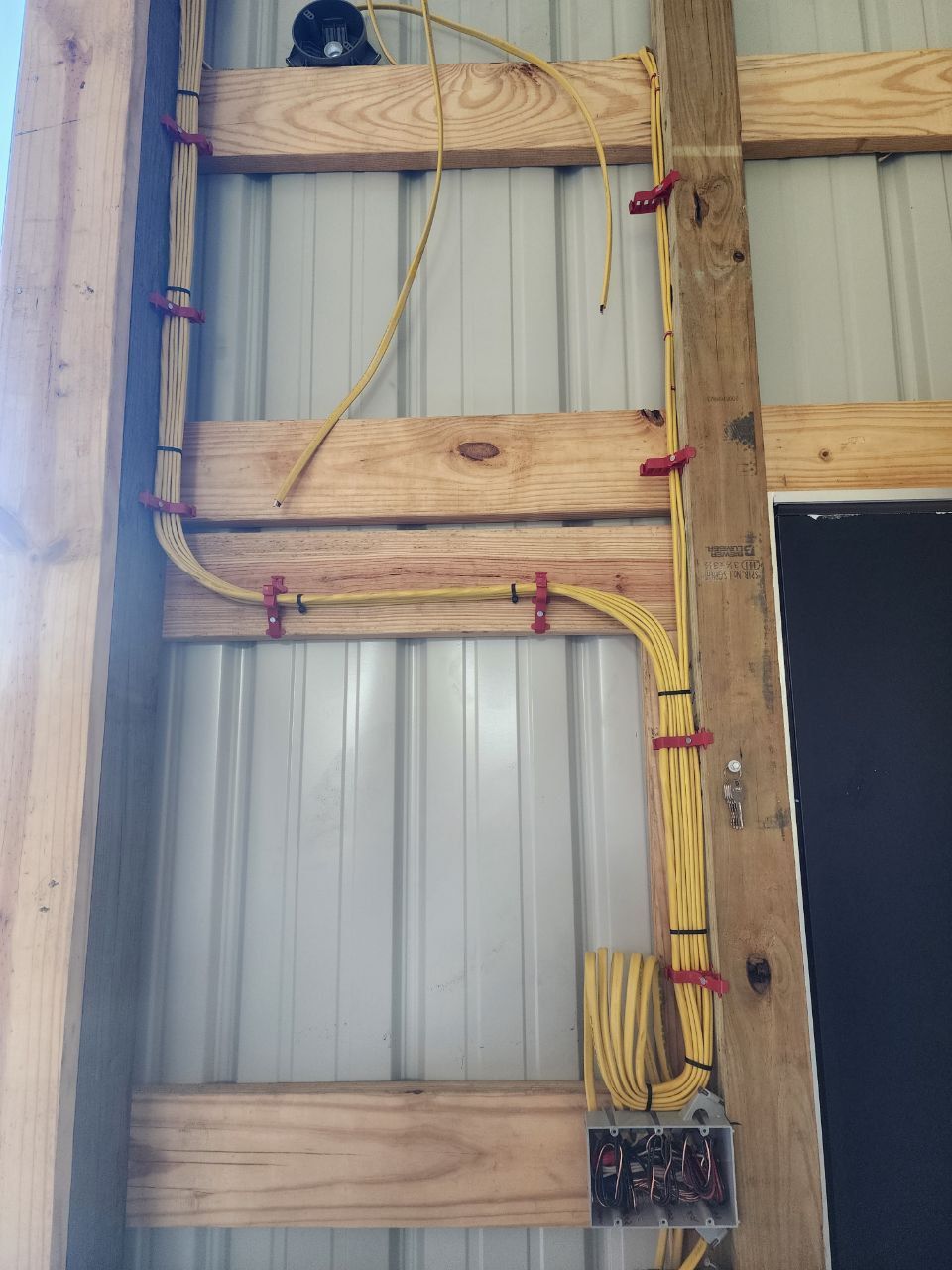 Electrical wiring attached to wooden beams with red clips in a metal-walled building.