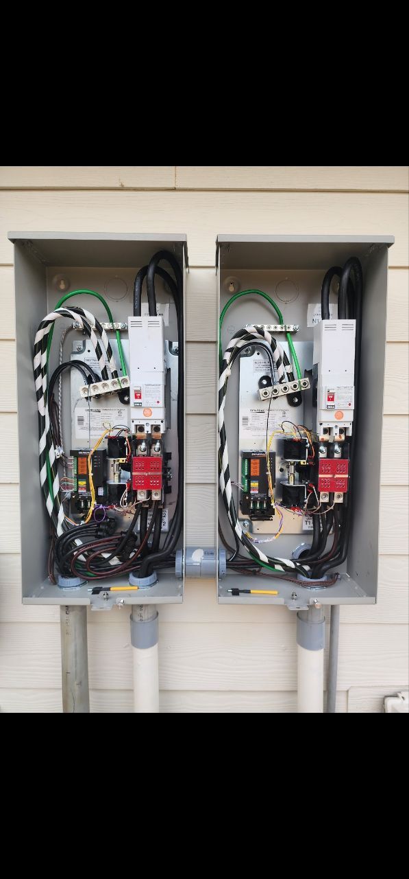Two electrical panels, grey boxes, mounted on a wall. Exposed wiring, conduit, and circuit breakers visible.