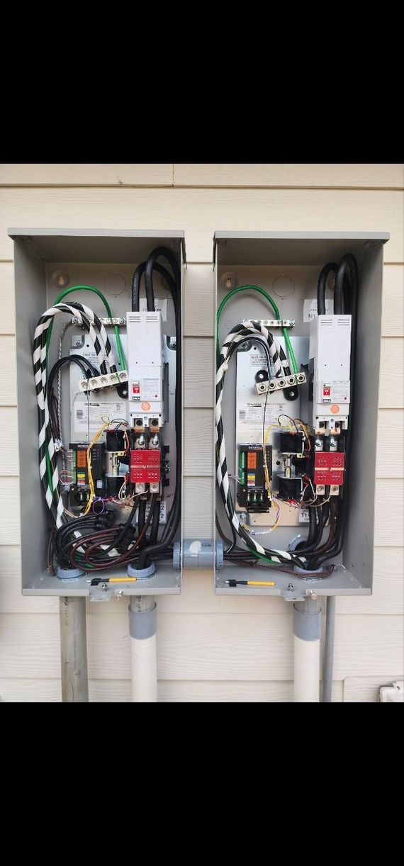 Two electrical panels, grey boxes, mounted on a wall. Exposed wiring, conduit, and circuit breakers visible.