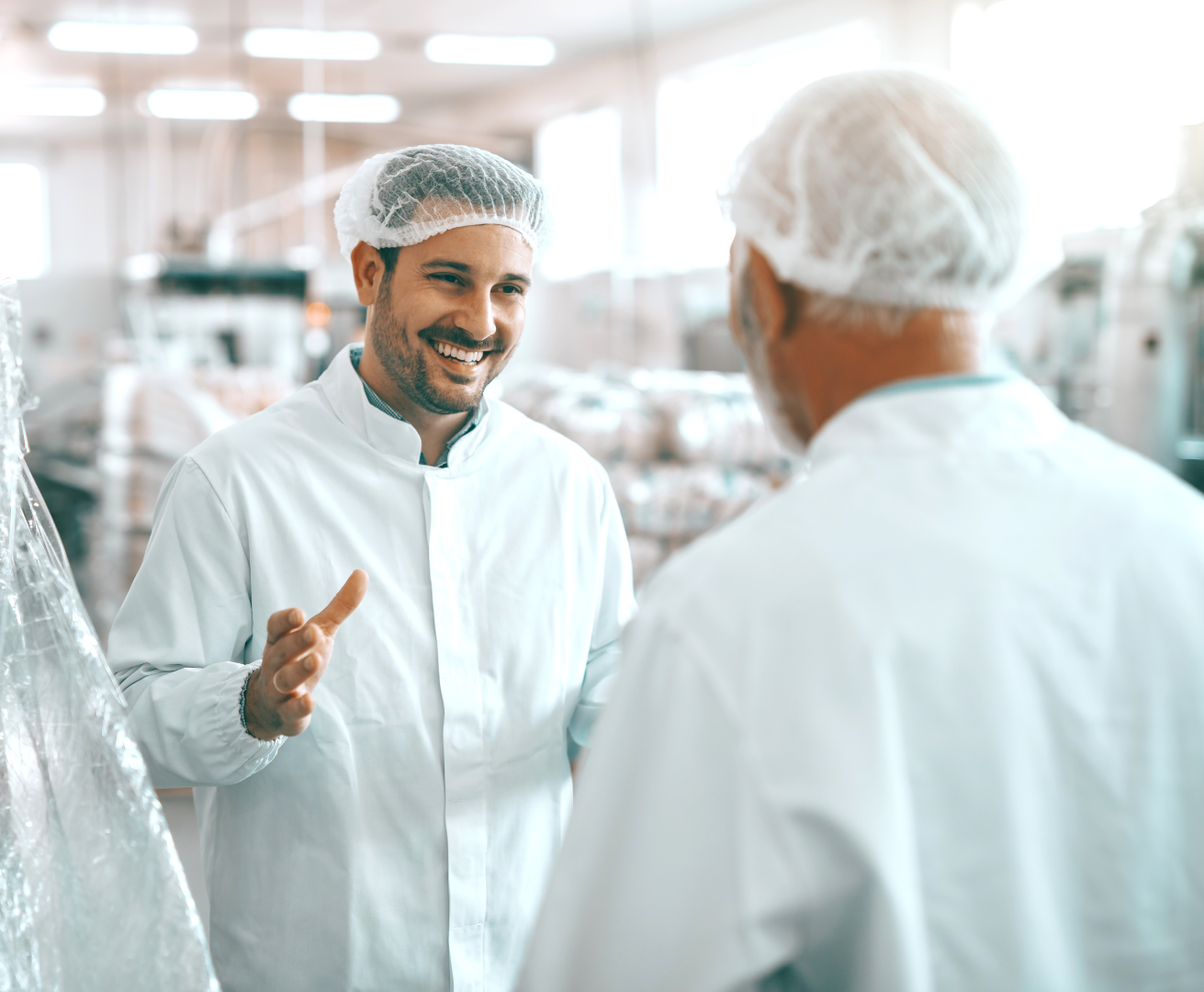 Two men in white coats and hair nets are talking to each other in a factory.