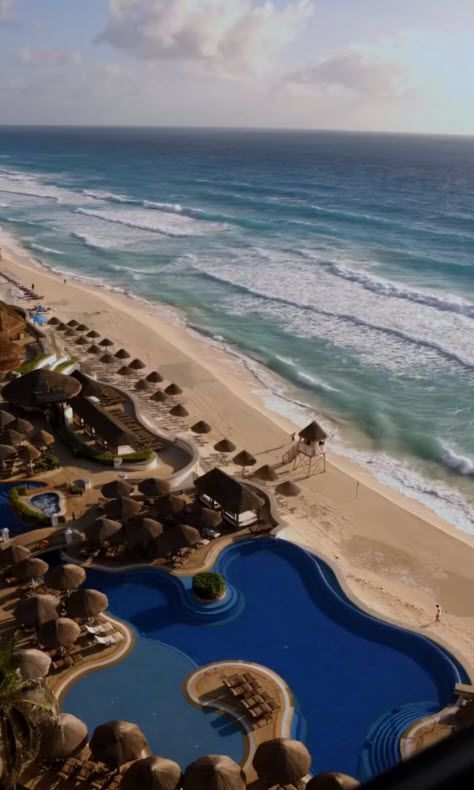 Beach resort view with ocean waves, sandy shore, palm-thatched umbrellas, and a blue pool.