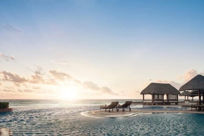 Infinity pool overlooks the ocean at sunrise; two lounge chairs, thatched-roof cabanas.