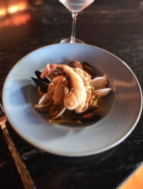 Seafood pasta dish in a blue bowl, with shrimp, mussels, and clams. A glass of wine is behind.