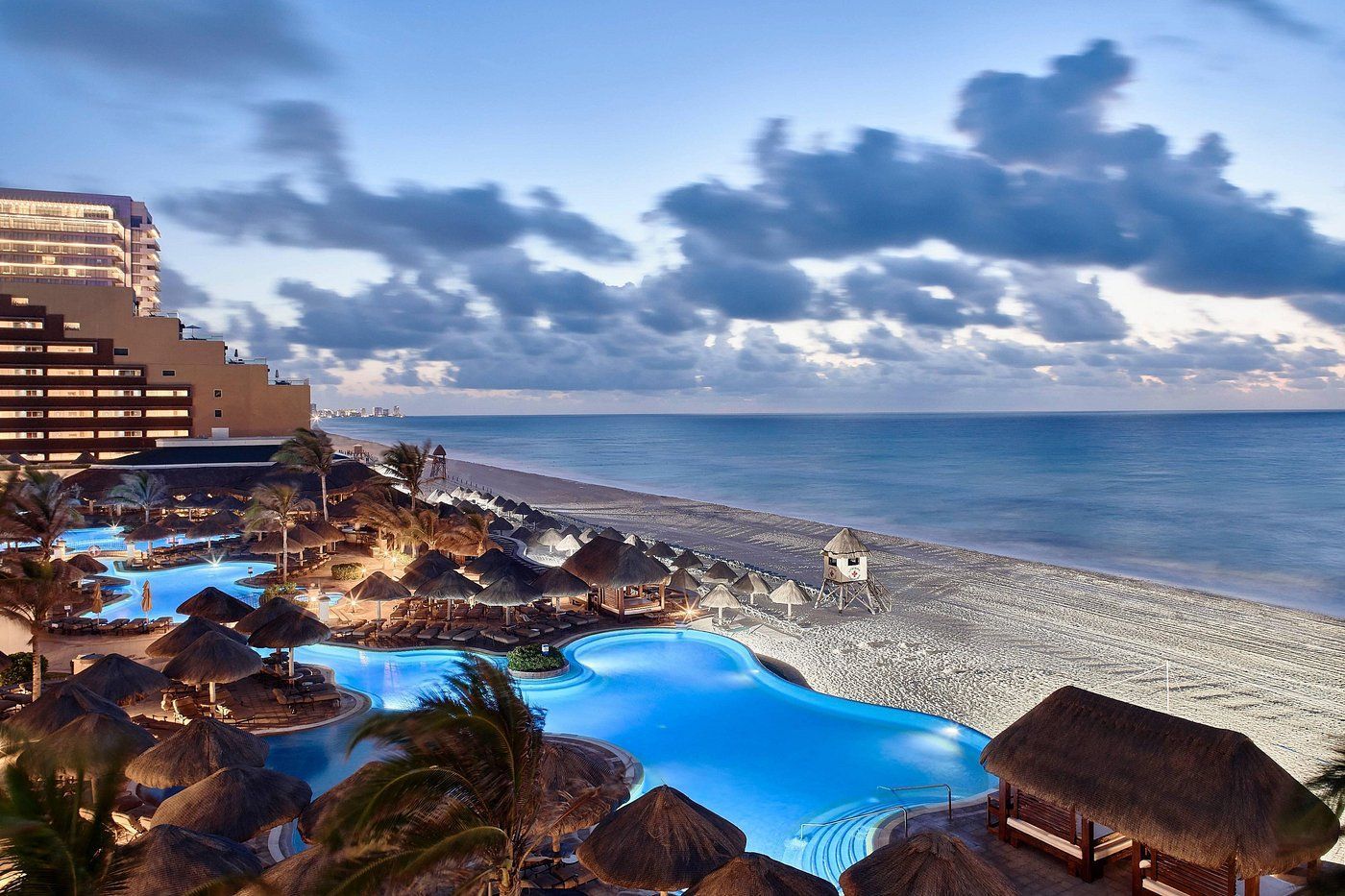 Resort pools and beach at dusk; ocean, thatched roofs, hotel.