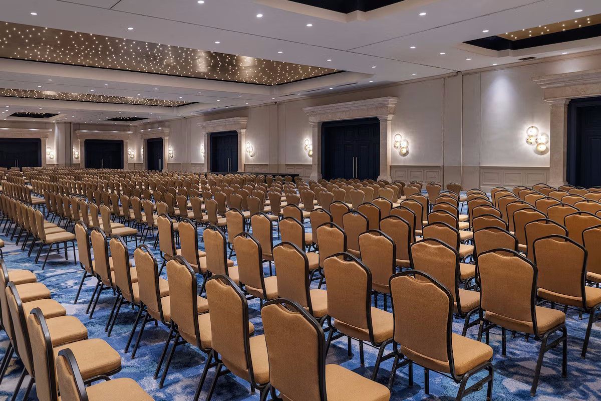 Empty auditorium with rows of tan chairs, blue carpet, and decorative ceiling lights.