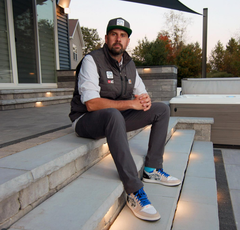 Man in vest, cap, and sneakers sitting on outdoor stone steps next to a hot tub.
