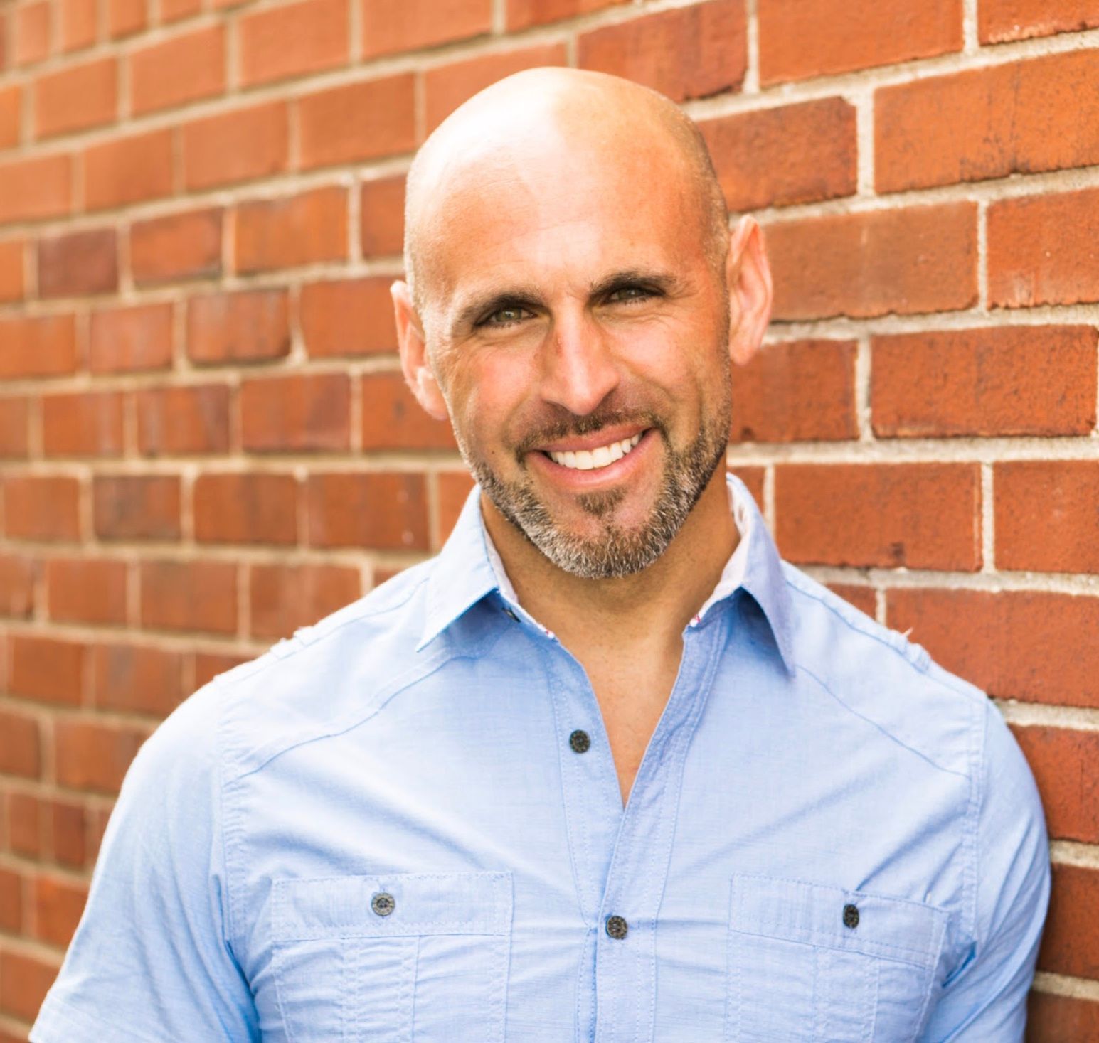 Bald man with a graying beard smiles, wearing a blue button-down shirt, leaning against a brick wall.