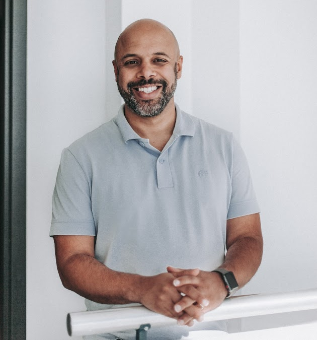 Man with a bald head and beard smiling, wearing a light gray polo shirt, leaning on a white railing.