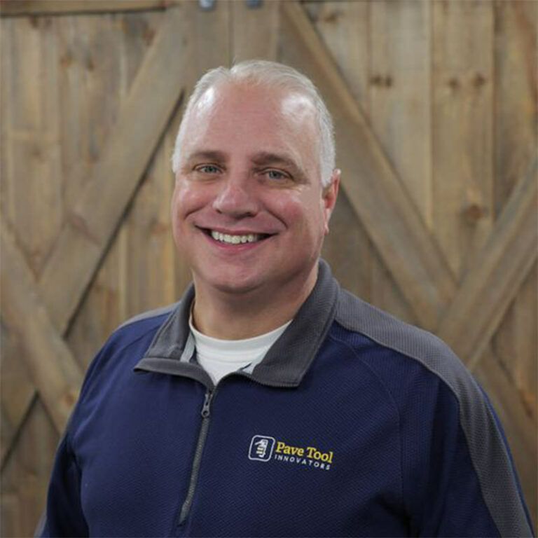 Man smiling in a blue and gray jacket with a company logo in front of a wooden background.