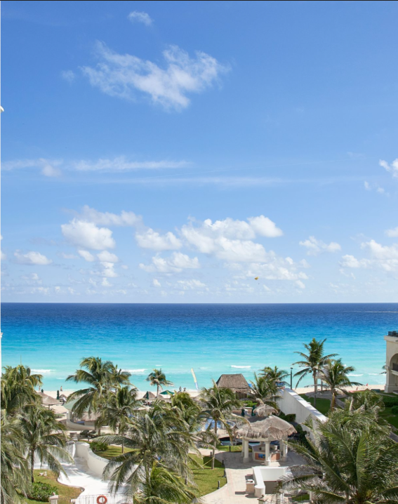 Bright blue ocean and sky over a beach with palm trees and a resort, under a partly cloudy sky.