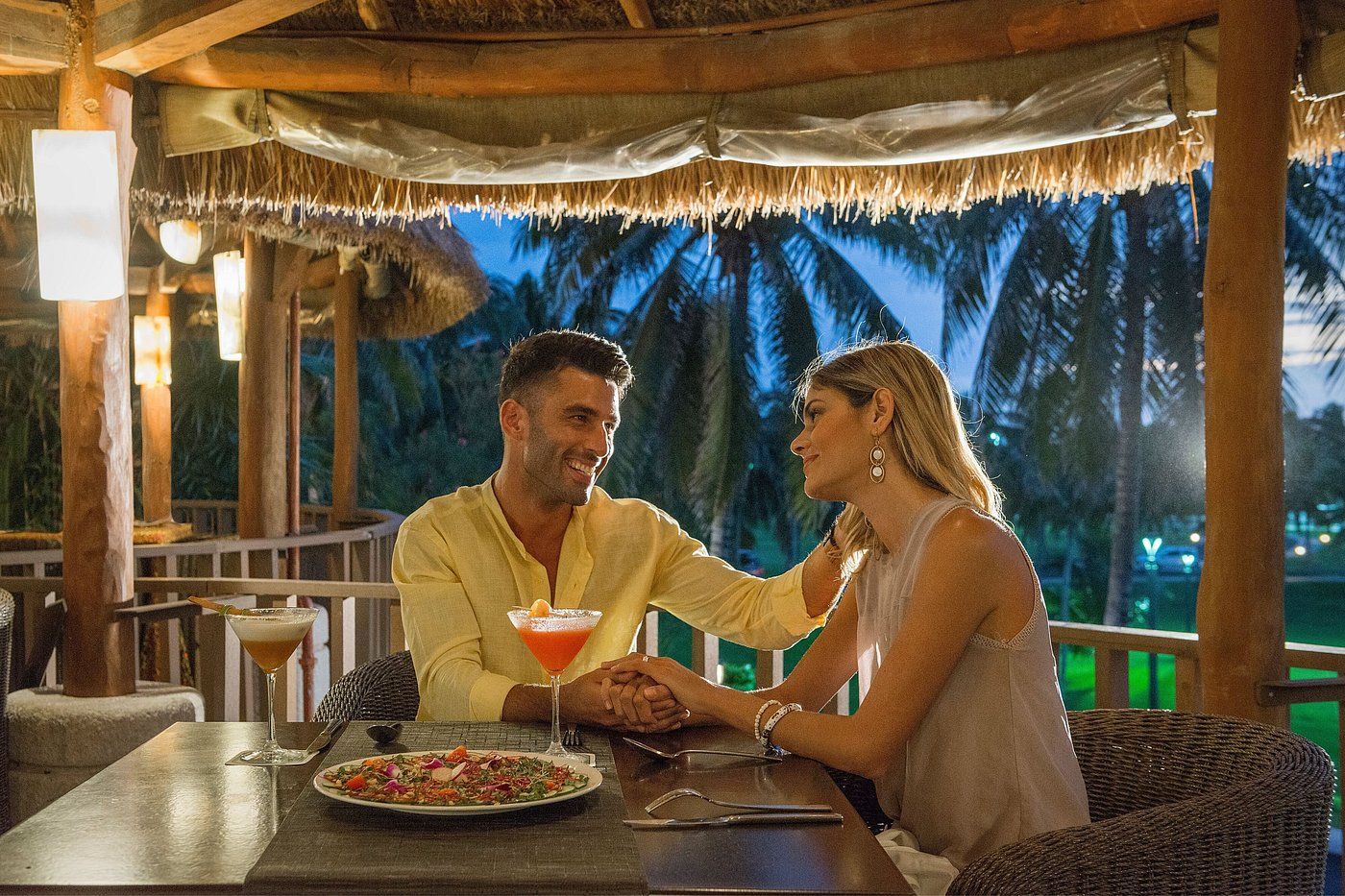 Couple at outdoor restaurant, holding hands, smiling. Palm trees and drinks visible.