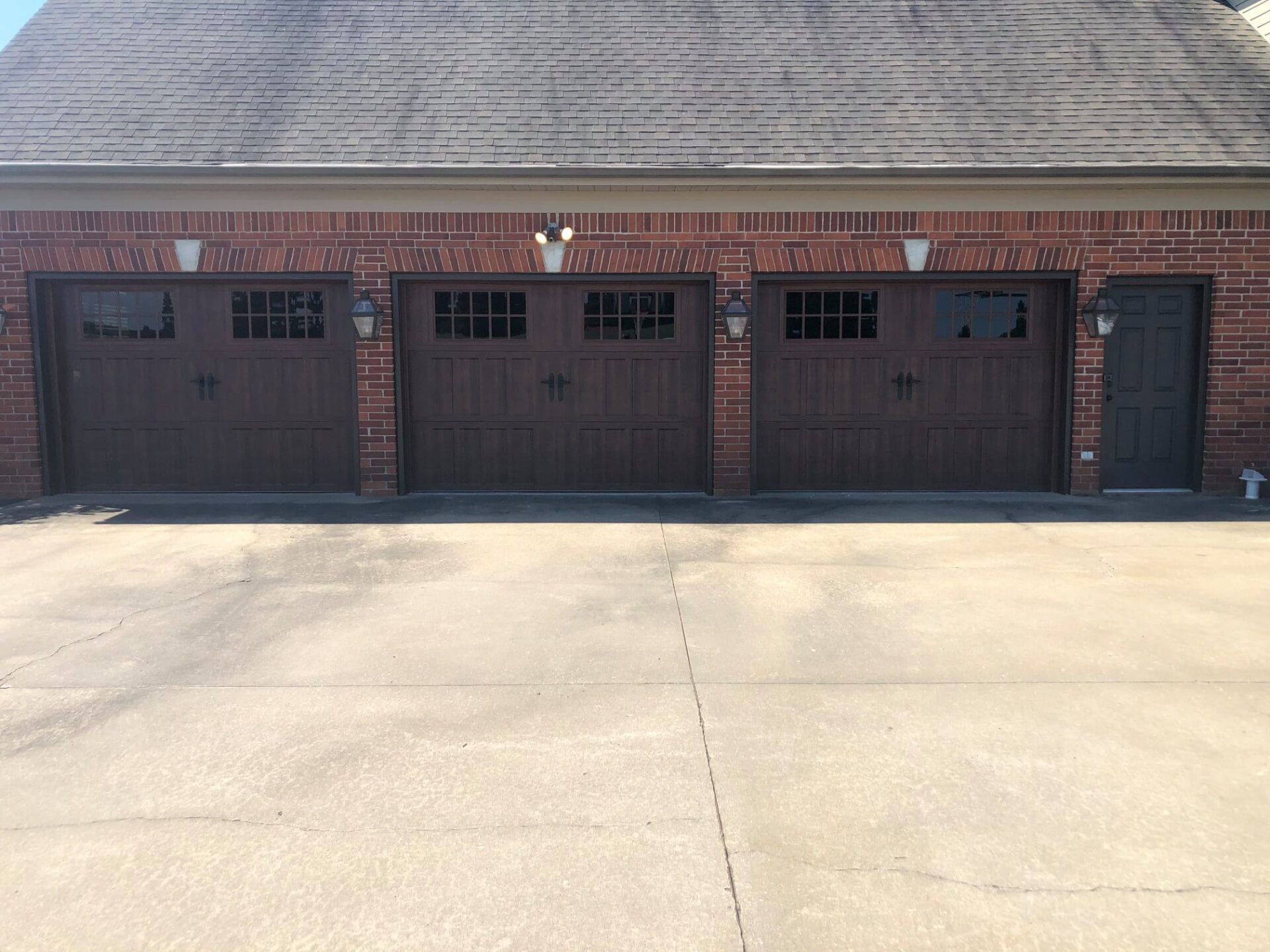 Three brown garage doors with window panes and a side door on a brick building, with a paved driveway.
