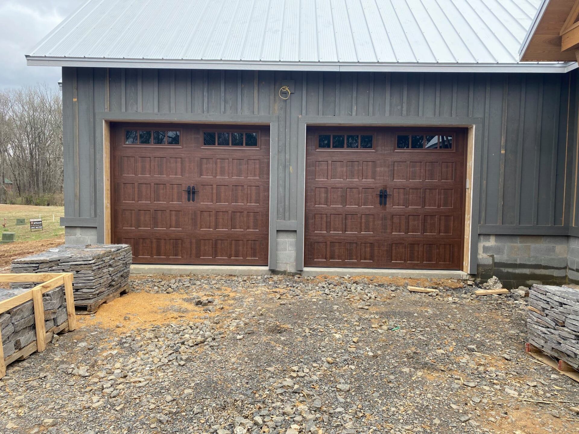 Two brown garage doors on a gray building with a metal roof. Construction materials are in the foreground.