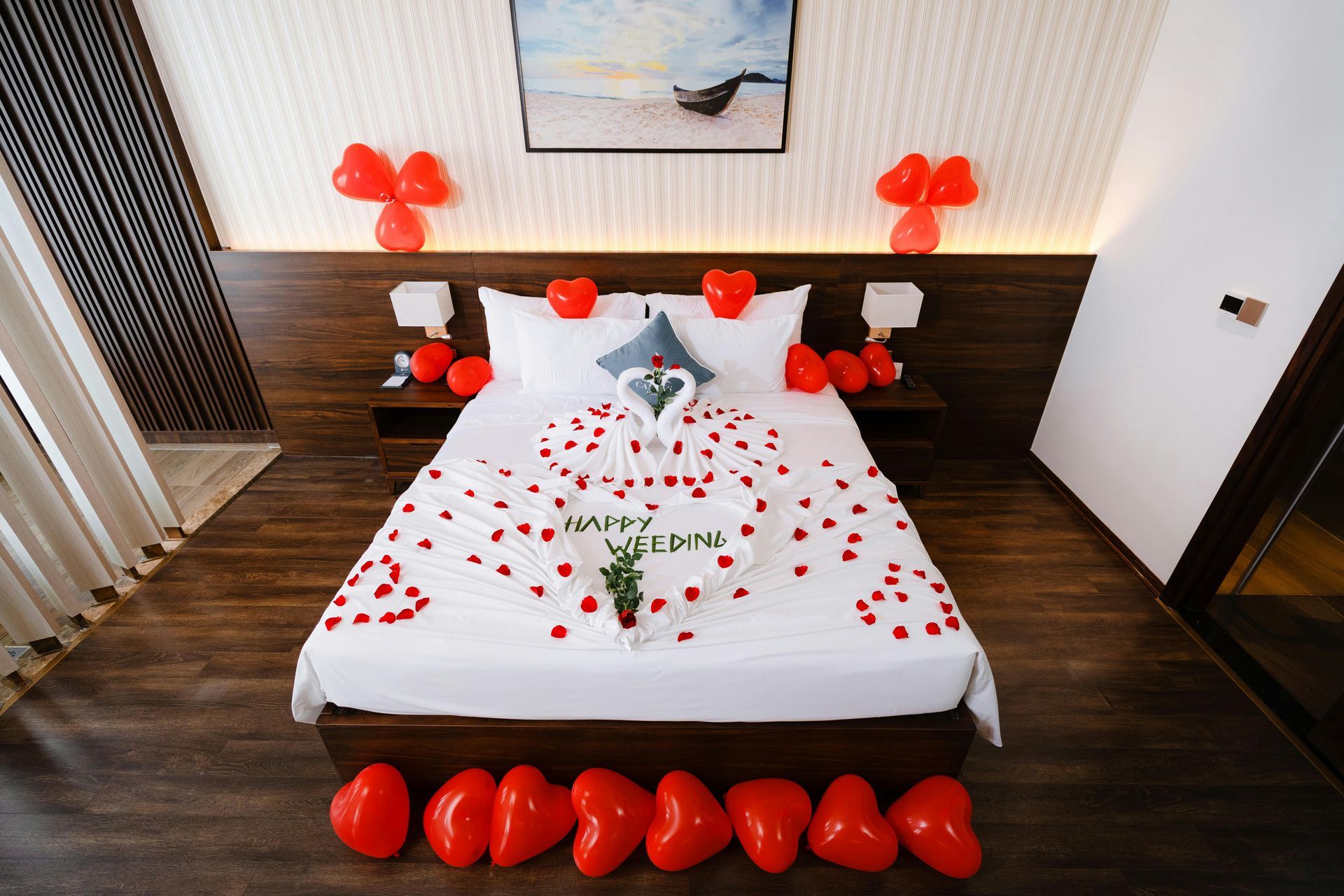 A romantic hotel bed decorated with red rose petals, heart-shaped balloon clusters, and a towel swan centerpiece.