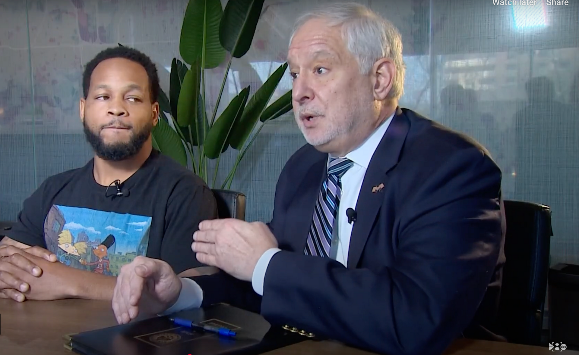 Two men, one in black, the other in a suit, in a room with bookshelves and an American flag, one handing a check.