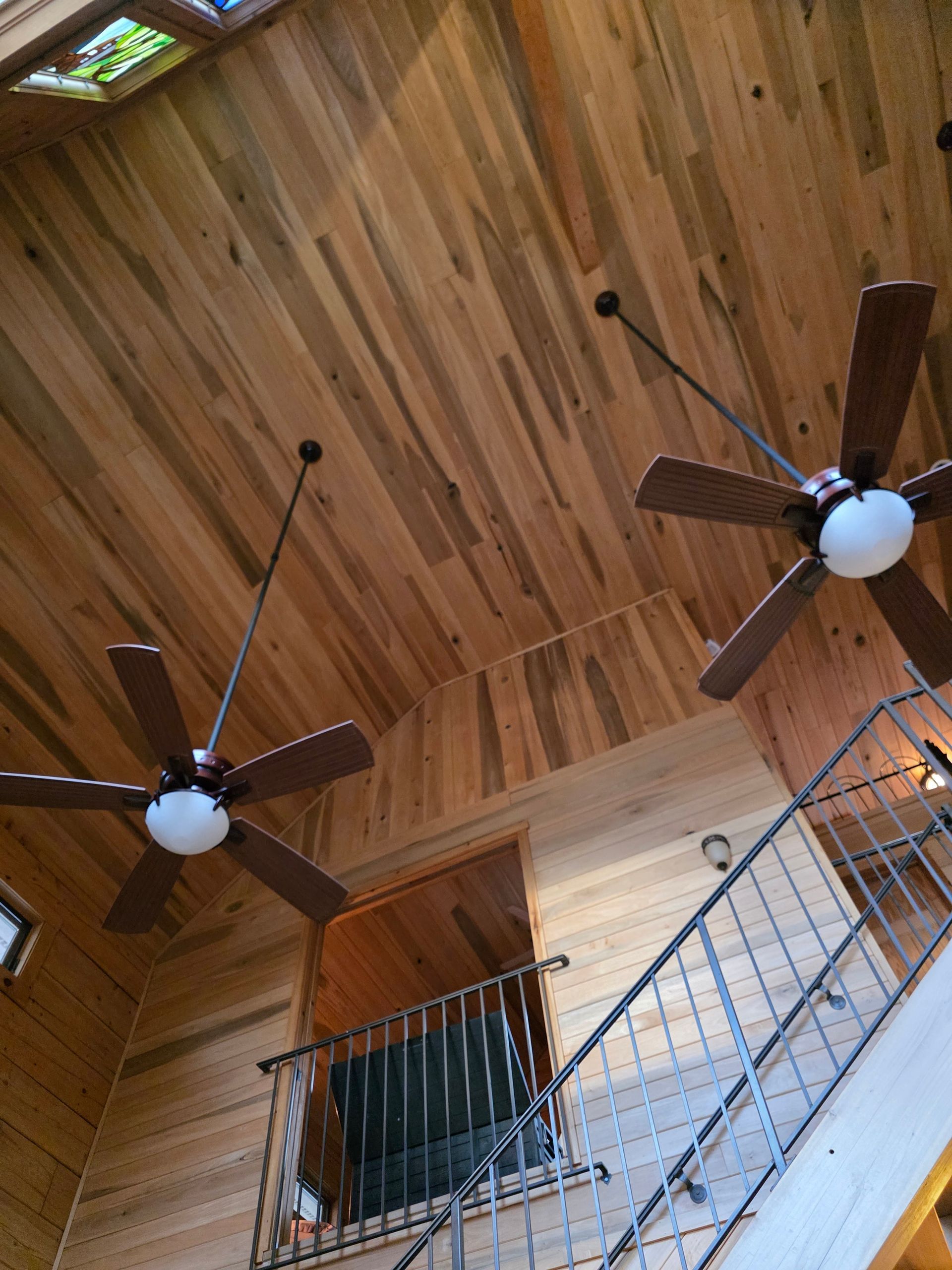 Wooden ceiling with two ceiling fans, stairs with metal railing, and a window.