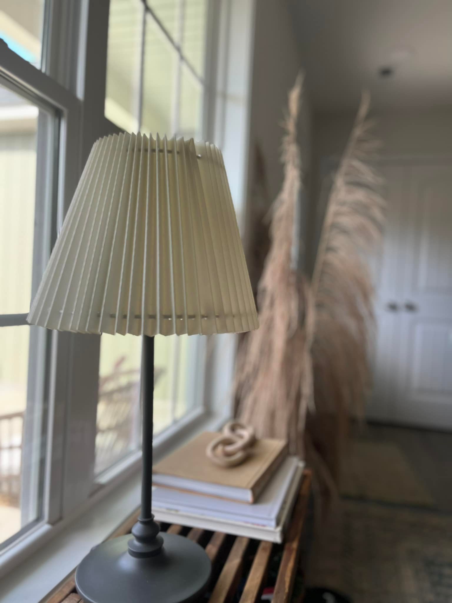 A lamp with a cream pleated shade sits on books by a window with dried pampas grass.