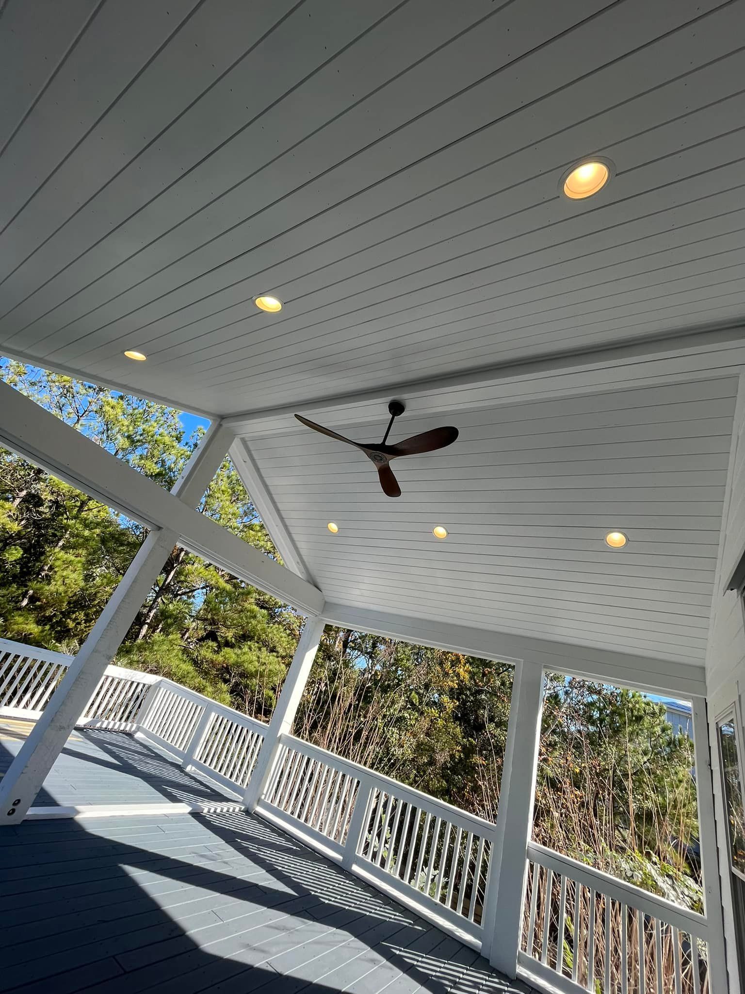Covered porch with white railings, ceiling fan, recessed lights, and view of trees.