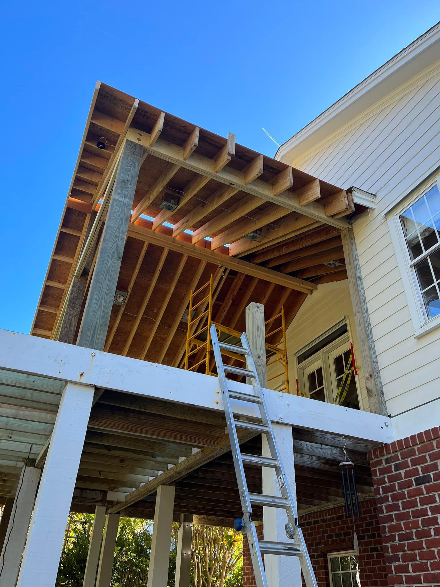 Construction of an outdoor wooden roof addition to a white house with a red brick base, blue sky background.