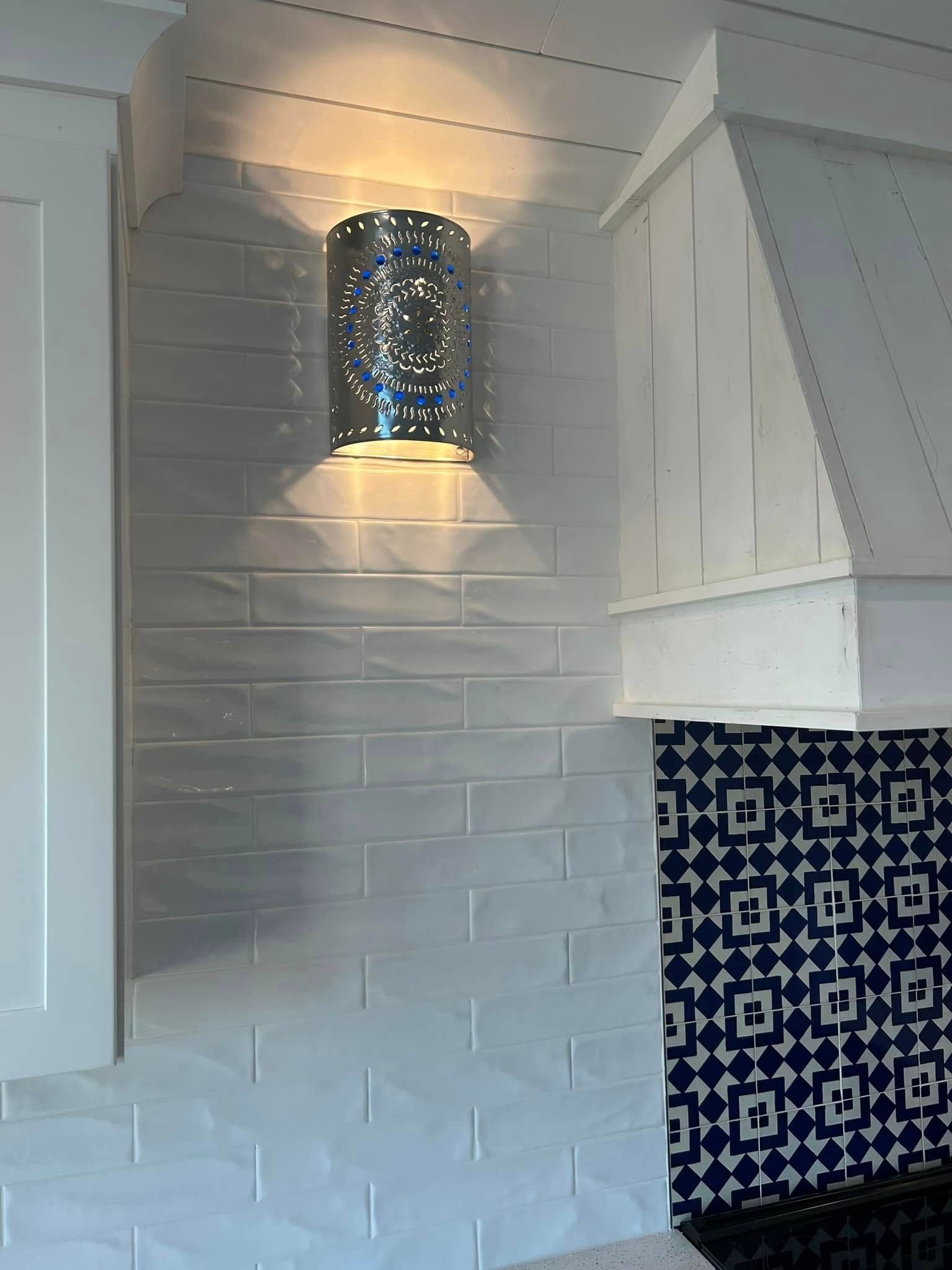 Silver wall sconce with perforated design emits light on white tiled backsplash.