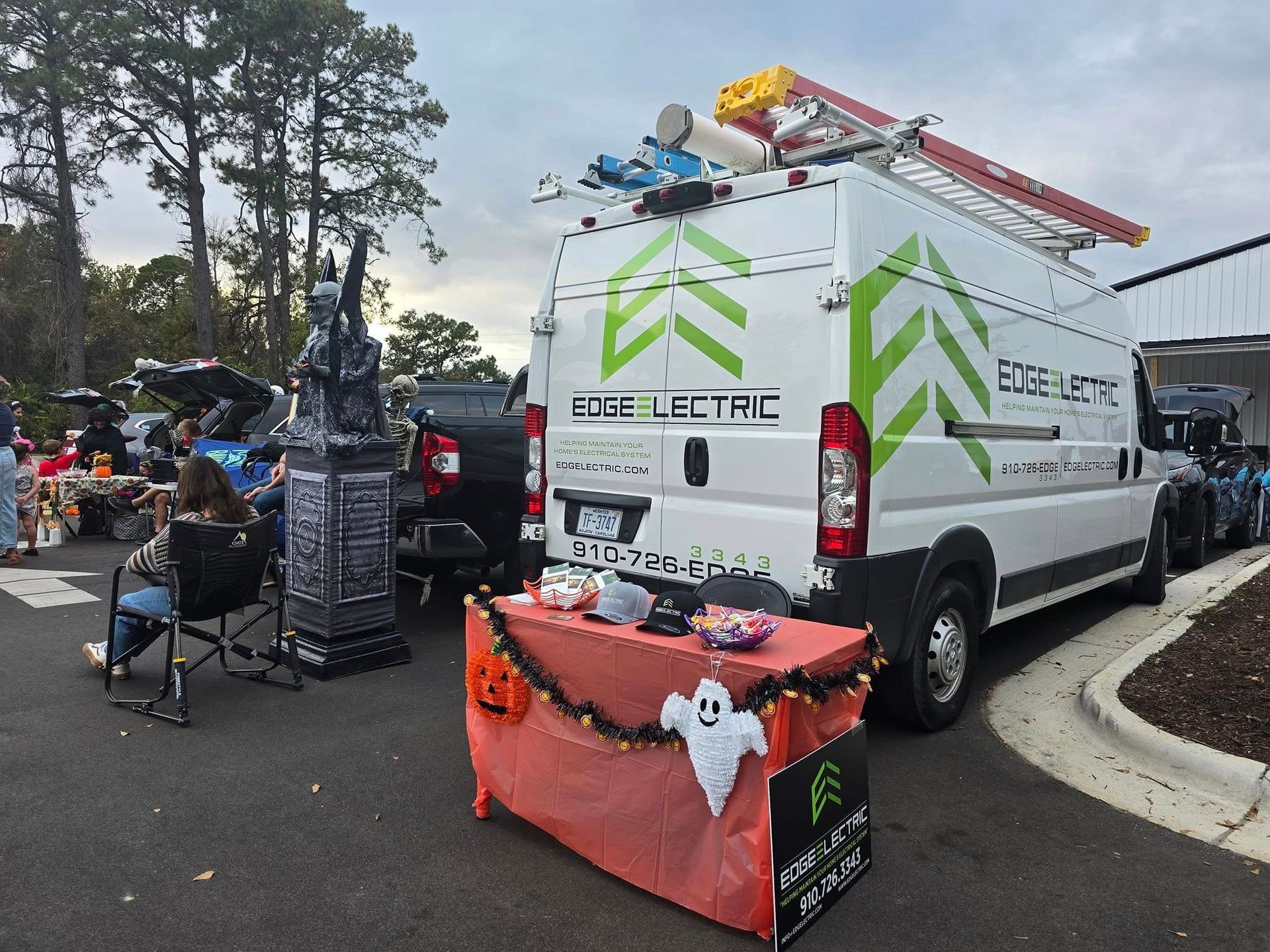 White van with green logo at a Halloween event; treats on a table with decorations, person in a chair.