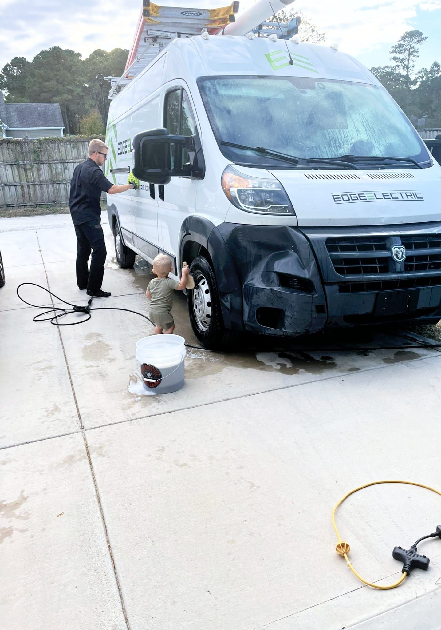 A man and small child wash a white van together on a concrete surface.