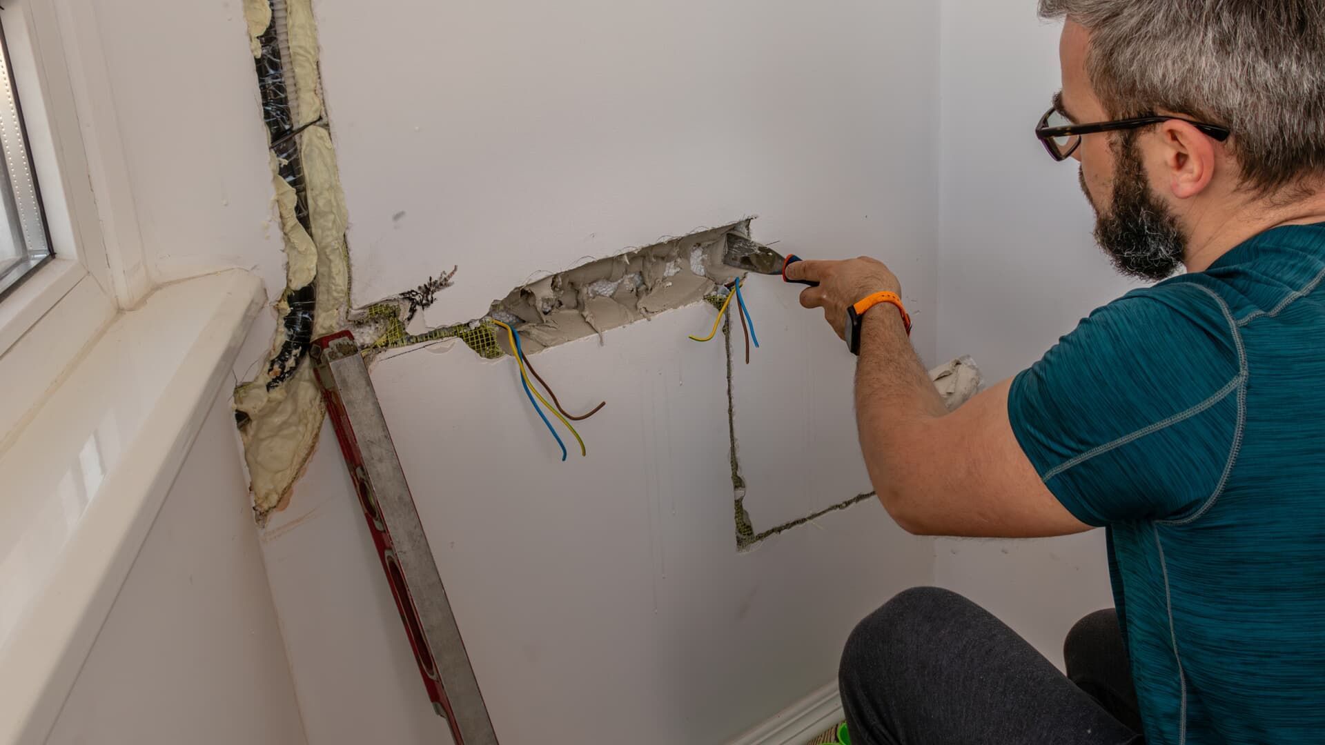 Man working on a wall, removing material. Wires exposed. White wall with window.
