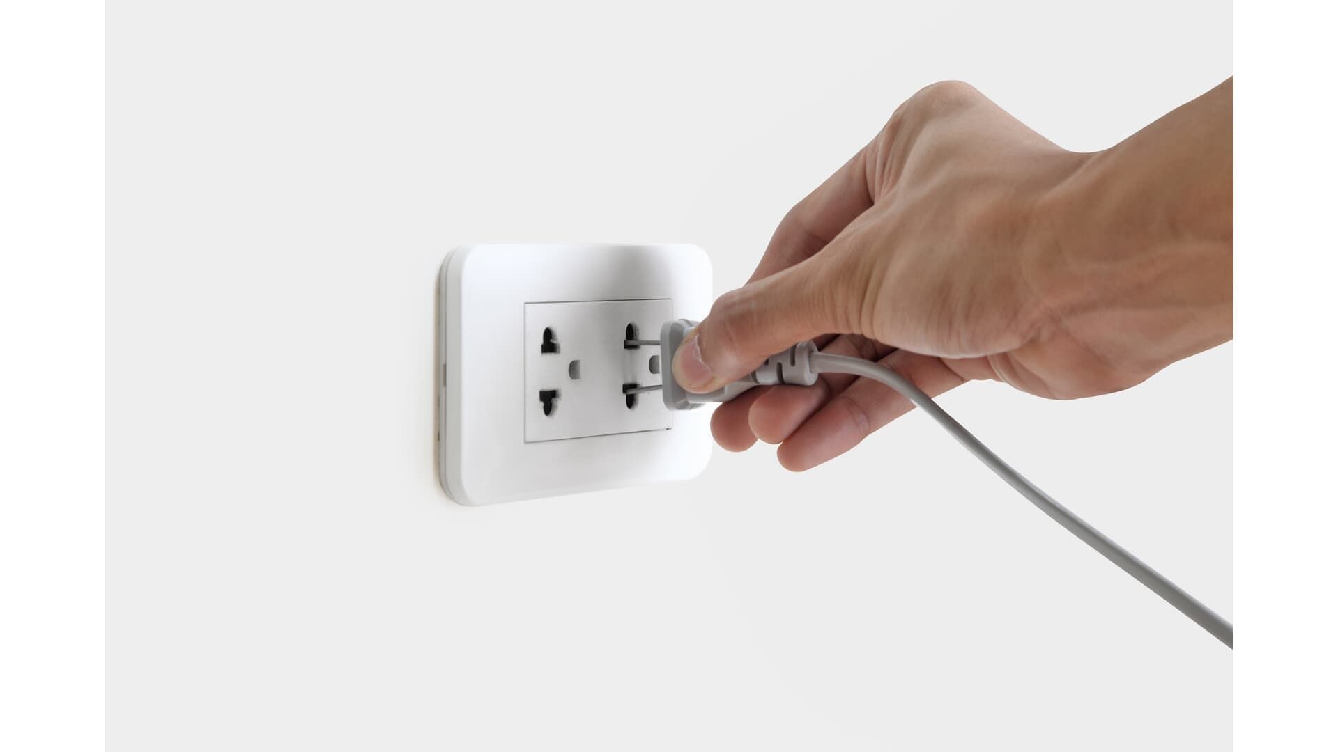 Hand plugging a gray electrical cord into a white wall outlet.