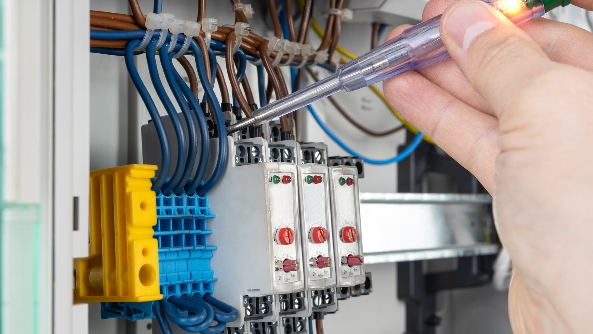 Hand testing electrical wires in a control panel with a glowing voltage tester.