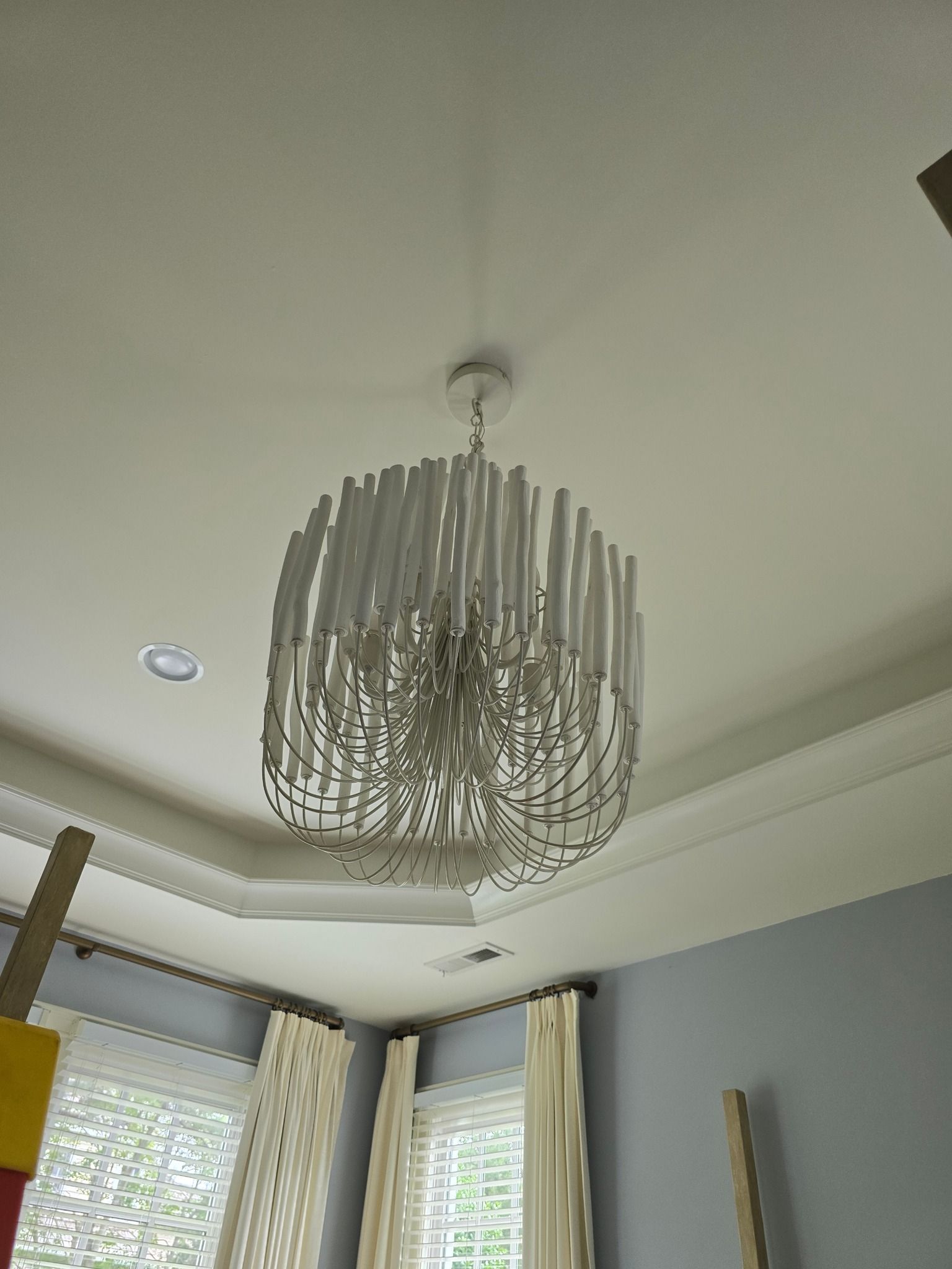 White chandelier hanging from a white ceiling in a room with a window and curtains.
