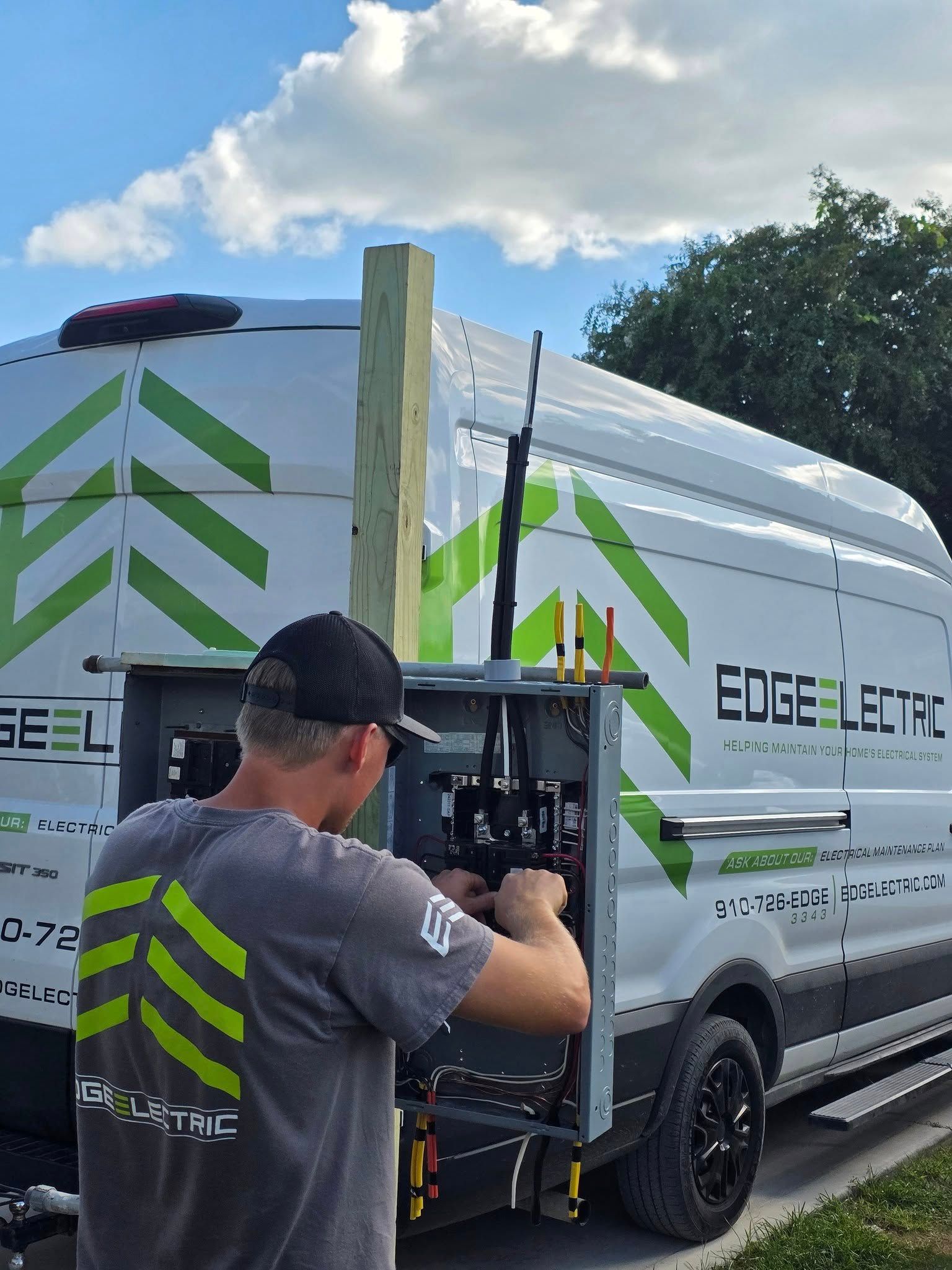 Electrician working on a gray electrical box on the back of a van. The van is white with green accents, blue sky.