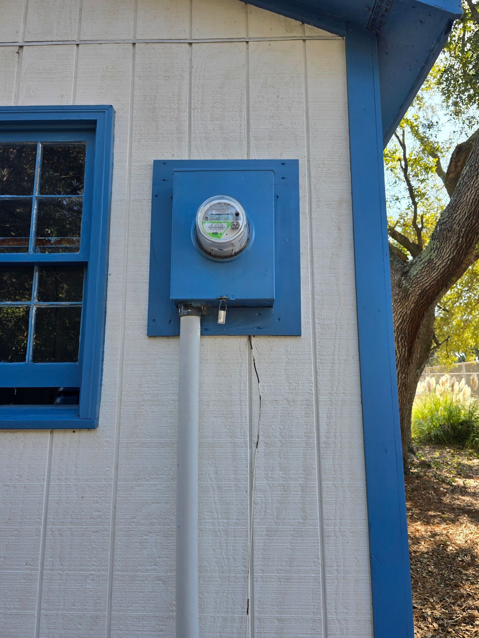 Blue electrical meter box on a white building, with a window to the left and a gray conduit below.