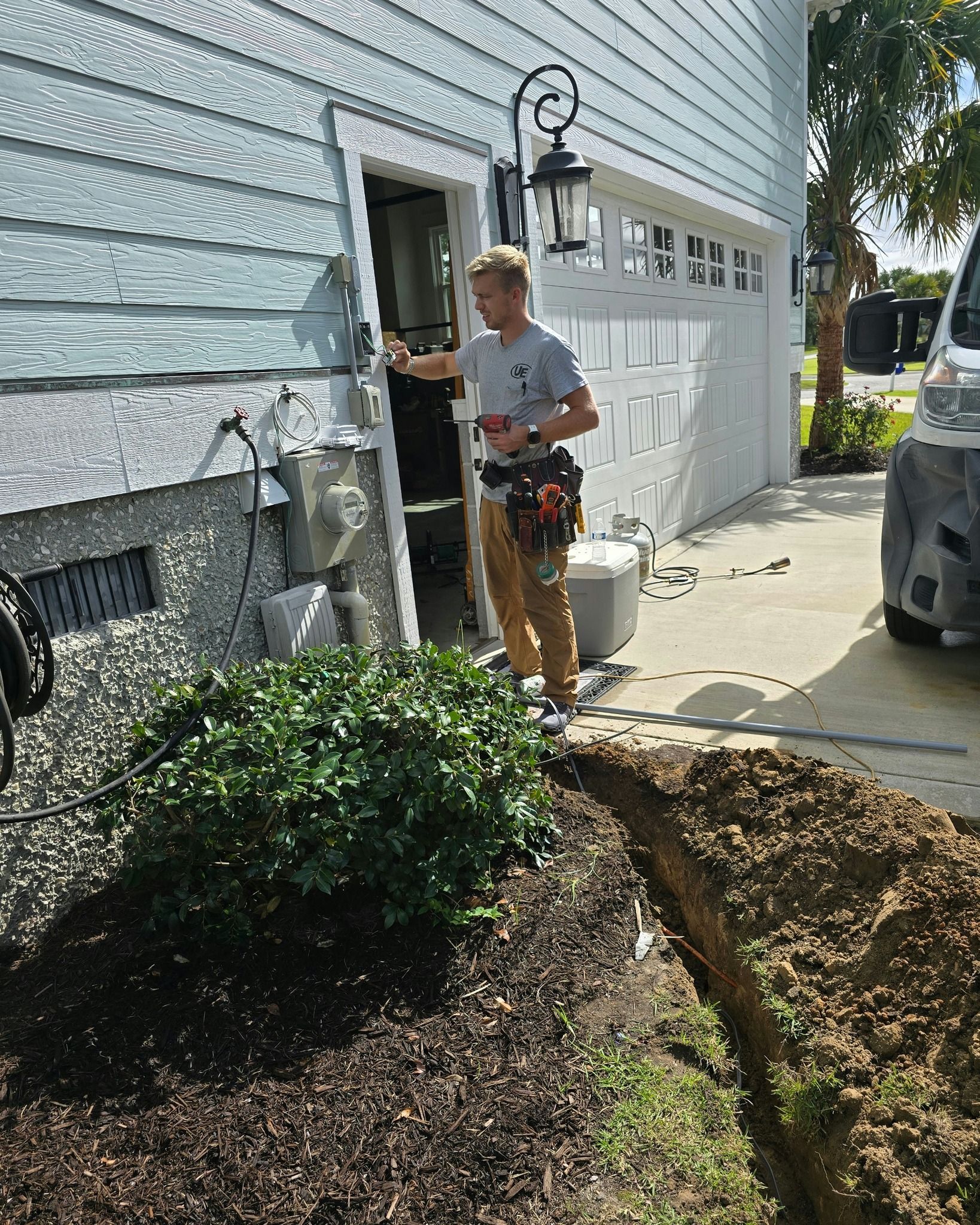Person in work clothes standing near open garage door, trench in foreground.