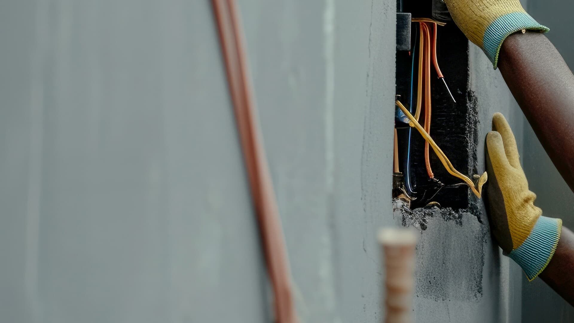 Hands wearing yellow gloves work on exposed electrical wires within a gray wall.