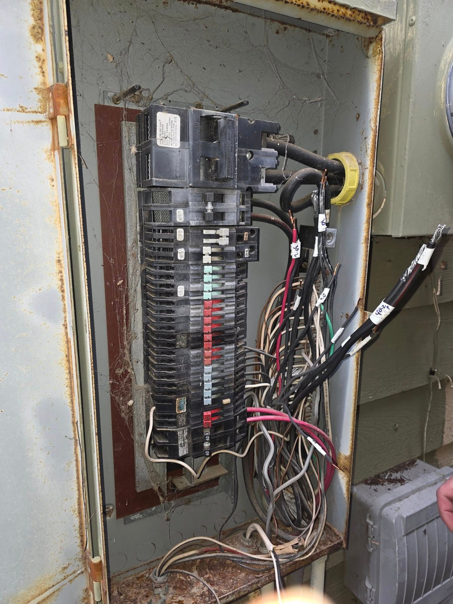 Electrical panel with exposed wires and breakers inside a rusted metal enclosure.